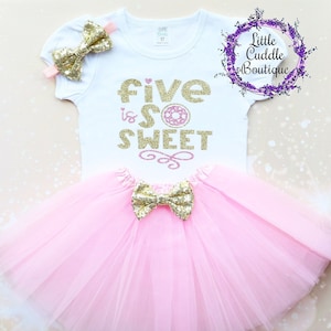 May include: A pink tulle tutu with a gold glitter bow. A white shirt with gold glitter text that says "five is so sweet" with a pink donut graphic.