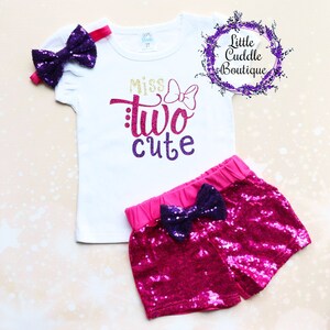 May include: A white t-shirt with pink and purple glitter lettering that says "Miss Two Cute" with a pink bow. The shirt is paired with pink sequined shorts with a purple bow.