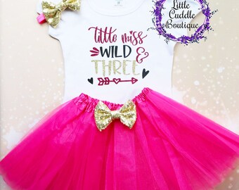May include: A white t-shirt with the text "little miss WILD & THREE" in gold glitter and maroon lettering. The shirt is paired with a pink tulle skirt and two gold glitter bows.