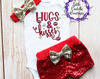 May include: A red and gold baby outfit with a white bodysuit that says "Hugs & Kisses" in red glitter. The outfit also includes a red headband with a gold bow and red sequined shorts with a gold bow.