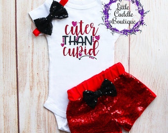 May include: A white bodysuit with the text "cuter than cupid" in pink glitter lettering and red hearts. The bodysuit is paired with red sequin shorts with a black bow.