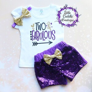 May include: A white t-shirt with the text "TWO & FABULOUS" in black and purple lettering with a black arrow pointing to the right. The shirt is paired with purple sequined shorts with a gold bow.