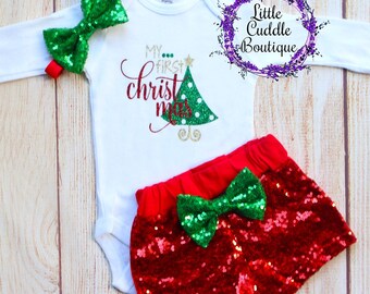 May include: A white long-sleeved bodysuit with the text "My first christmas" and a green and gold glitter Christmas tree. A red and green sequined shorts with a green bow. A green sequined bow headband.
