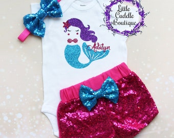 Peut inclure: Une tenue pour bébé en trois pièces avec un motif de sirène. La tenue comprend un body blanc avec une sirène turquoise aux cheveux violets, un short en sequins roses avec un nœud turquoise et un bandeau en sequins turquoise avec un nœud rose.