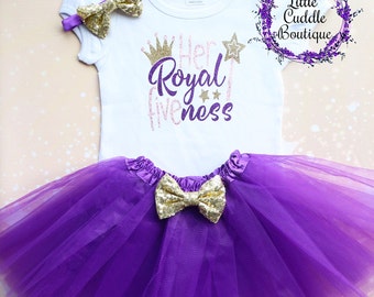 Peut inclure: Un body blanc avec le texte "Her Royal Highness" en paillettes dorées et violettes. Le body est associé à une jupe en tulle violette et un nœud doré.