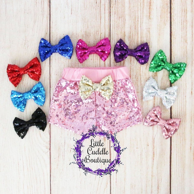 Op de afbeelding: Roze sequin shorts met een gouden strik aan de voorkant. De shorts worden omringd door een verscheidenheid aan kleurrijke strikken, waaronder rood, blauw, paars, groen en zilver.