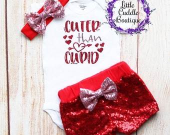 May include: A white bodysuit with the text "Cuter than Cupid" in red glitter with a red heart and arrow. The bodysuit is paired with red sequin shorts with a pink bow. A pink and red headband with a bow is included.