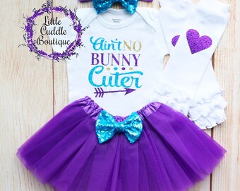 May include: A baby girl outfit with a white bodysuit that says "Ain't No Bunny Cuter" in gold and teal glitter, a purple tulle skirt, a teal sequin bow headband, and white leg warmers with a purple glitter heart.