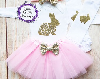 May include: A baby girl's Easter outfit with a white bodysuit featuring a gold glitter bunny and the text "my first easter", a pink tulle tutu with a gold glitter bow, a white headband with a gold glitter bow, and white socks with gold glitter bunny designs.