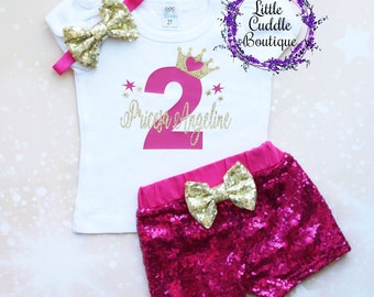 Peut inclure: Un t-shirt blanc avec un numéro deux rose et le texte "Princess Angeline" en paillettes dorées. Le t-shirt est associé à un short rose à paillettes avec un nœud doré.