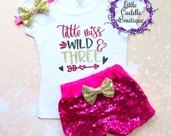 May include: A white toddler t-shirt with the text "Little Miss Wild & Three" in pink, black, and gold glitter. The set includes matching pink sequin shorts and a gold sequin bow headband. A gold sequin bow is also on the shorts.