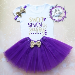 Puede incluir: Una camiseta blanca con texto de purpurina dorada que dice "Sweet Seven & Sassy" con un coraz&oacute;n morado. La camiseta se combina con un tut&uacute; de tul morado y una diadema de lazo de purpurina dorada.