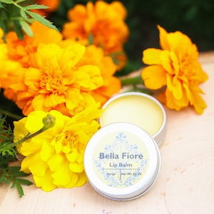 May include: A small silver tin of Bella Fiore lip balm with a mango scent. The tin is open and the lip balm is visible. The tin is surrounded by yellow marigold flowers.