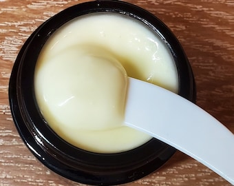 May include: A close-up shot of a jar of cream with a white spatula. The cream is a pale yellow color and has a smooth, creamy texture. The jar is black with a wide opening.