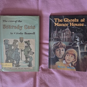 May include: Two vintage children's books. "The Case of the Scaredy Cats" by Crosby Bonsall features a group of children. "The Ghosts at Manor House" has a spooky illustration of a house and children. Both books are on a pink background.