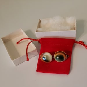 May include: A pair of cufflinks with a round, iridescent shell design. The cufflinks are in a red velvet pouch and a white box.