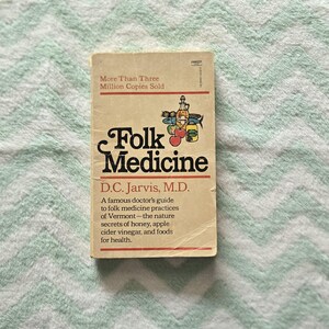 May include: A paperback book titled Folk Medicine by D.C. Jarvis, M.D. The beige cover features a small illustration of a bottle, fish, and fruit, with the text More Than Three Million Copies Sold and a description of Vermont folk medicine practices.