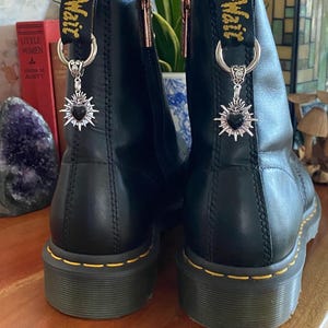 May include: A pair of black leather ankle boots featuring yellow stitching along the soles and black heel pull tabs with yellow text. Each boot is adorned with a silver-toned metal charm shaped like a sacred heart with a black center stone.
