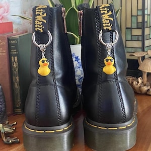 May include: A pair of black leather ankle boots featuring yellow stitching along the sole and a black heel pull tab with yellow text. Each boot is adorned with a silver-toned metal ring holding a small yellow rubber duck charm wearing sunglasses.