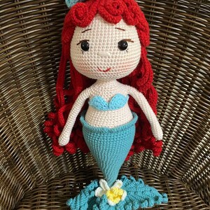 Pode incluir: Boneca de croch&ecirc; de sereia com cabelo ruivo, top de concha azul e cauda turquesa. A boneca tem um rosto branco com olhos pretos e um pequeno sorriso. Uma coroa de flores brancas e azuis est&aacute; na cabe&ccedil;a.