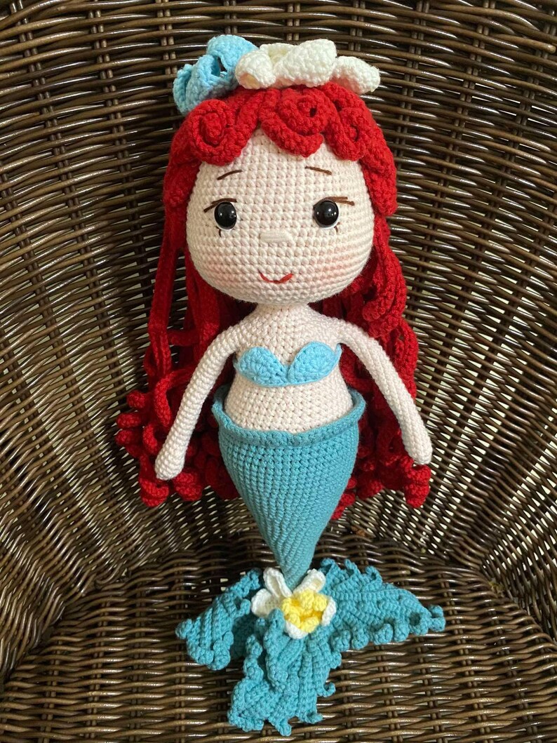 Pode incluir: Boneca de croch&ecirc; de sereia com cabelo ruivo, top de concha azul e cauda turquesa. A boneca tem um rosto branco com olhos pretos e um pequeno sorriso. Uma coroa de flores brancas e azuis est&aacute; na cabe&ccedil;a.