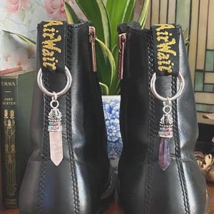 May include: A pair of black leather boots featuring black heel pull tabs with yellow AirWair embroidery. Each boot has a silver-toned metal ring attached to the pull tab, holding a dangling crystal pendant with a silver-toned decorative cap.
