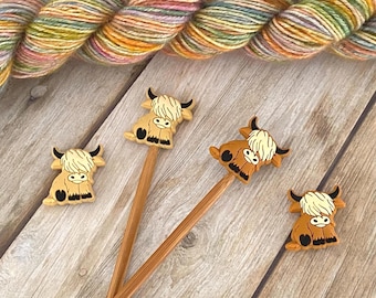 May include: Two bamboo knitting needles topped with brown and cream Highland cow figures, accompanied by two matching Highland cow buttons. A skein of variegated yarn in shades of green, yellow, pink, and blue is positioned above the needles.
