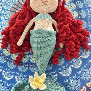 Pode incluir: Boneca de sereia de croch&ecirc; feita &agrave; m&atilde;o com cabelo encaracolado vermelho, top azul claro e cauda turquesa. A boneca tem rosto e bra&ccedil;os cor creme e uma flor branca e amarela na cauda. A boneca est&aacute; sobre uma superf&iacute;cie azul estampada.