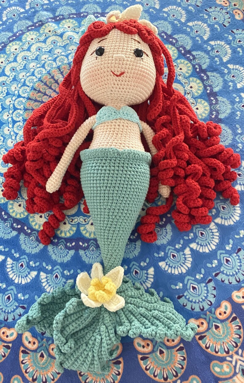 Pode incluir: Boneca de sereia de croch&ecirc; feita &agrave; m&atilde;o com cabelo encaracolado vermelho, top azul claro e cauda turquesa. A boneca tem rosto e bra&ccedil;os cor creme e uma flor branca e amarela na cauda. A boneca est&aacute; sobre uma superf&iacute;cie azul estampada.