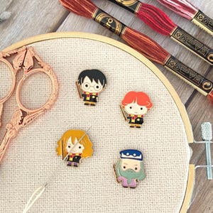 May include: Four enamel pins depicting cartoon characters with wands are arranged on a beige embroidery hoop. A pair of rose gold scissors with ornate handles and three skeins of red and brown embroidery floss are positioned nearby.