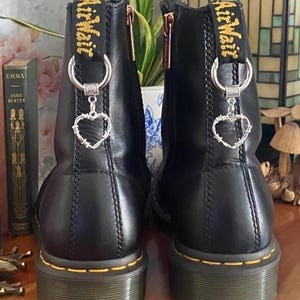 May include: A pair of black leather ankle boots featuring yellow stitching along the sole and a yellow heel pull tab with black text. Each boot is adorned with a silver-toned metal ring and a dangling heart-shaped charm made of barbed wire.