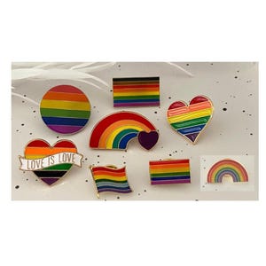 May include: A collection of seven enamel pins featuring rainbow designs, including a circle, a rectangular flag, a heart, a rainbow with a small heart, a heart with a banner reading LOVE IS LOVE, a waving flag, and a small rainbow arch.