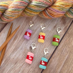 May include: Five stitch markers shaped like miniature yarn balls in purple, yellow, green, pink, and light blue, each with a red band featuring a white character face and a silver lobster clasp, accompanied by two wooden knitting needles.