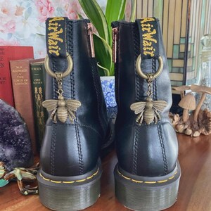 May include: A pair of black leather ankle boots featuring yellow stitching along the sole, a side zipper, and a black heel pull tab with yellow AirWair text. Each boot is adorned with a metallic bronze bee charm hanging from a circular ring.