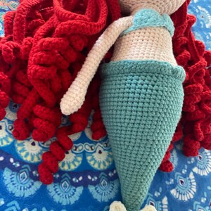 Pode incluir: Boneca de sereia de croch&ecirc; feita &agrave; m&atilde;o, com cabelo encaracolado vermelho, um top azul claro e uma cauda turquesa. A boneca tem um corpo e bra&ccedil;os cor de creme e um detalhe floral na base da cauda. A sereia est&aacute; sobre um fundo azul padronizado.