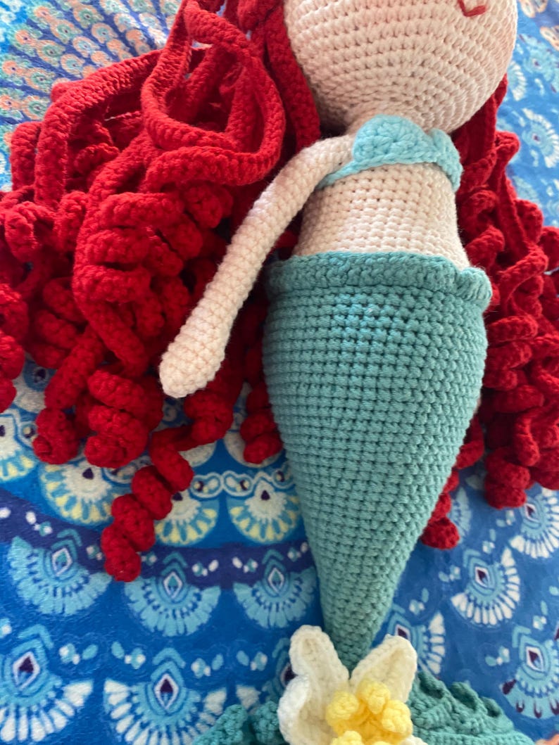 Pode incluir: Boneca de sereia de croch&ecirc; feita &agrave; m&atilde;o, com cabelo encaracolado vermelho, um top azul claro e uma cauda turquesa. A boneca tem um corpo e bra&ccedil;os cor de creme e um detalhe floral na base da cauda. A sereia est&aacute; sobre um fundo azul padronizado.