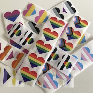 May include: A selection of heart-shaped stickers displaying various LGBTQ+ pride flags. The stickers showcase rainbow, bisexual, transgender, and other pride flag designs. The stickers are presented on a white background.