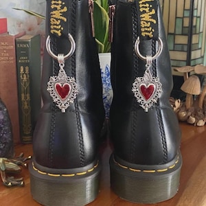 May include: A pair of black leather ankle boots featuring yellow stitching along the soles and black heel loops with yellow text. Each boot is adorned with a silver-toned metal charm shaped like a sacred heart with a red center, attached to a ring.