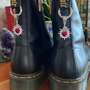 May include: A pair of black leather ankle boots featuring yellow stitching along the sole and a black heel loop with yellow text. Each boot is adorned with a silver-toned metal charm shaped like a sunburst heart with a red center.