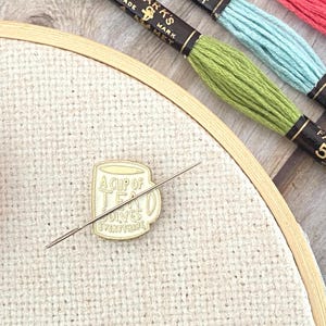 May include: A cream-colored enamel needle minder shaped like a mug with the text A CUP OF TEA SOLVES EVERYTHING, featuring a silver needle resting across it, placed on a beige embroidery hoop with three skeins of embroidery floss in green, blue, and red.