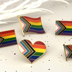 May include: A selection of gold-coloured enamel pins displaying the Progress Pride flag. The pins are in diverse shapes, including flags and a heart, with a rainbow of colours and a gold border.