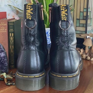May include: A pair of black leather ankle boots featuring yellow stitching along the sole, side zippers, and black heel pull tabs with the yellow text AirWair. Each boot is adorned with a silver-toned ring and a black sunburst heart charm.