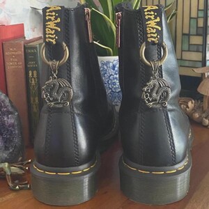 May include: A pair of black leather ankle boots featuring yellow stitching along the sole, a side zipper, and a yellow heel pull tab with the text AirWair. Each boot is adorned with a circular bronze-toned dragon charm attached to the heel loop.