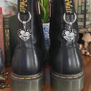 May include: A pair of black leather ankle boots featuring yellow stitching along the soles and black heel loops with yellow text. Each boot is adorned with a silver-toned metal heart-shaped charm depicting two skeletons kissing, attached to a ring.