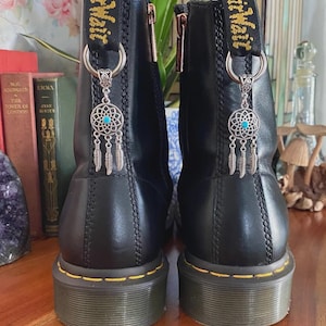 May include: A pair of black leather boots featuring yellow stitching along the sole and a black heel pull tab with yellow text. Each boot is adorned with a silver-toned metal dreamcatcher charm featuring a small blue stone and three feathers.