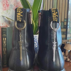 May include: A pair of black leather ankle boots featuring a vertical zipper on the side and a black heel pull tab with yellow text. Each boot is adorned with a metal ring attached to the pull tab, holding a dangling sword-shaped charm.