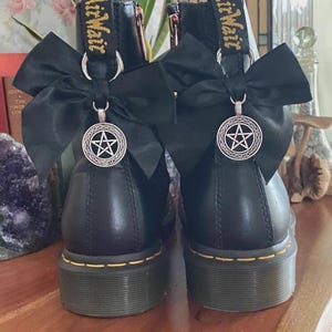 May include: A pair of black leather boots featuring yellow stitching along the sole and a black fabric heel loop with the text AirWair. Each boot is adorned with a large black satin bow and a silver-toned circular pendant featuring a pentagram.
