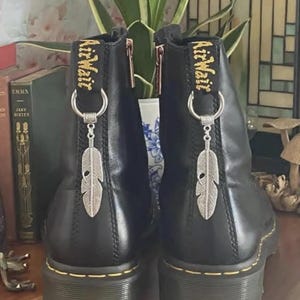 May include: A pair of black leather ankle boots featuring yellow stitching along the soles and black heel pull tabs with the yellow text AirWair. Each boot is adorned with a silver-toned metal ring and a dangling silver feather charm.