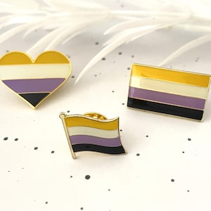 May include: Three enamel pins displaying the non-binary pride flag. One is heart-shaped, another rectangular, and the third a small flag. Each pin features yellow, white, purple, and black stripes.