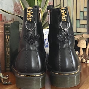 May include: A pair of black leather ankle boots featuring yellow stitching along the sole, a side zipper, and a black moth charm attached to a silver ring on the heel pull tab. The pull tab displays the yellow text AirWair in a script font.